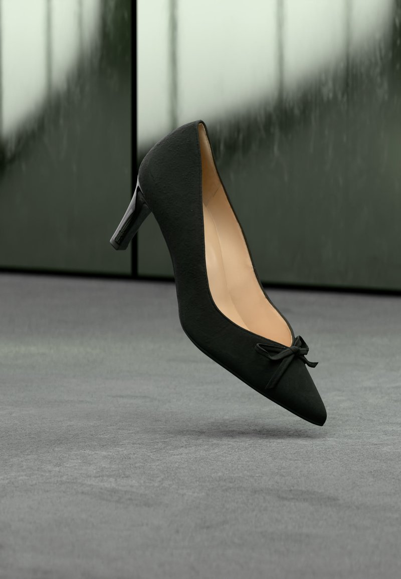 Chaussure à talon haut en daim noir avec bout pointu et petit nœud, flottant au-dessus d'un sol recouvert de moquette grise sur un fond flou.