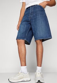 Persona vistiendo pantalones cortos de mezclilla azules hasta la rodilla, calcetines blancos hasta el tobillo y zapatillas Adidas de color blanco roto, de pie contra un fondo claro y liso.