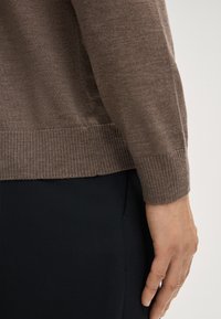 Braun gestrickter Pullover mit geripptem Saum und Bündchen, kombiniert mit schwarzen Stoffhosen. Fokus auf Textur und Farbkombination.