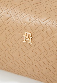Sac en cuir tan avec un motif géométrique en relief et un accent de logo doré. La texture est lisse avec une finition brillante.