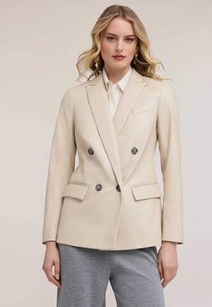 Kvinde med løst blondt hår iført gyldne øreringe, en beige dobbeltradet blazer over en hvid skjorte og grå bukser, stående mod en neutral baggrund.