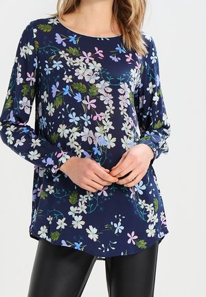 Femme portant une tunique bleu marine à manches longues avec un imprimé floral multicolore, associée à un pantalon noir, debout les mains jointes.