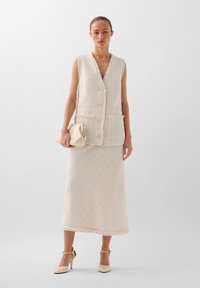 Gilet beige in tweed con scollatura a V, quattro bottoni e tasche frontali, abbinato a una lunga gonna coordinata. Entrambi presentano motivi testurizzati.