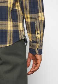 Camisa de franela a cuadros en azul marino y amarillo con cuello y puños abotonados. Presenta un dobladillo curvado y una tela texturizada.