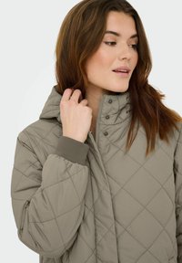 Prešit lahno sivo jakno z visokim ovratnikom, gumbi na pritrditev in rebrastimi zapestji. Dizajn ima vzorec diamantov in gladko teksturo.