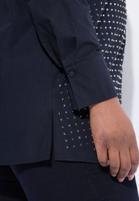 Dark navy shirt with long sleeves, featuring a row of silver studs along the side seam. Fabric appears smooth with a structured fit.
