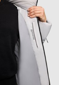 Hellgraue Jacke mit glatter Textur, Innentaschendetail und schwarzem Reißverschluss, von vorne betrachtet, mit einem schwarzen Pullover darunter.