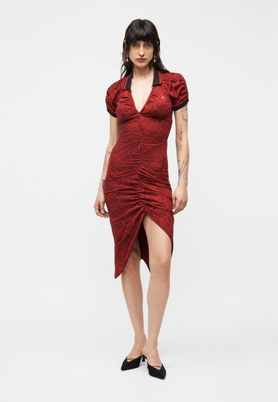 Γυναίκα με σκούρα μαλλιά, που φοράει εφαρμοστό κόκκινο φόρεμα με ρεβρά μαύρο γιακά και φουσκωτά μανίκια, στέκεται σε μαύρες μυτερές γόβες μπροστά σε λευκό φόντο.