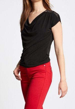 Mujer con una blusa negra de manga corta y cuello drapeado, y pantalones ajustados rojos, de pie con una mano cerca de la cadera.