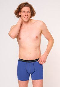 Boxer ajustados de hombre en color azul, hechos de una tela suave, con una cinturilla negra que lleva el logo, diseño ajustado y costuras en contraste en las aberturas de las piernas.