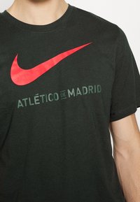 Detailní záběr osoby v tmavě zeleném tričku s červeným logem Nike ve tvaru háčku a textem "Atlético de Madrid" na hrudi.