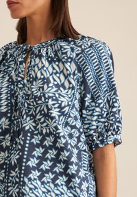 Blusa blu navy con motivi floreali in bianco e azzurro. Maniche a sbuffo con dettaglio a fiocco sul colletto. Tessuto morbido e liscio.