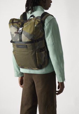 Kamerat bag i grønt og brunt stoff, med en fold-over topp, justerbare stropper og en frontlogo-patch.