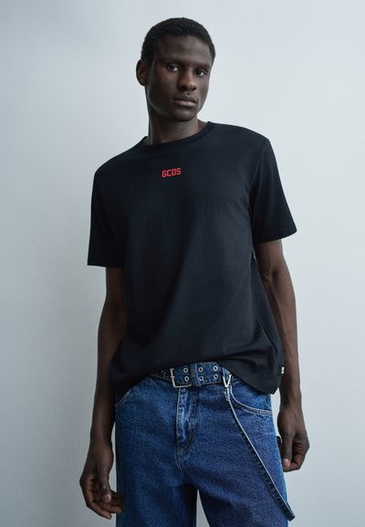 Άνδρας που φοράει μαύρο t-shirt της GCDS και μπλε τζιν με ζώνη και ιμάντα, στέκεται μπροστά σε μονόχρωμο ανοιχτό γκρι φόντο.