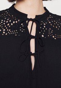 Blouse noire avec panneaux d'épaule en dentelle et trois nœuds attachés le long de l'encolure fendue devant, portée par une personne aux cheveux courts.