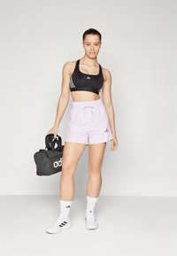 Top desportivo preto com riscas brancas, calções cor-de-rosa claro com cós elástico, meias brancas e mochila preta. Ténis brancos nos pés.