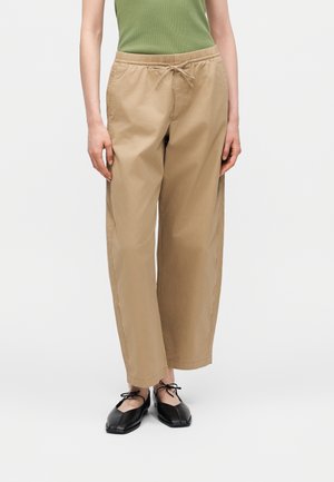 Άτομο που φοράει μπεζ παντελόνι με κορδόνι, ελιάς πράσινο αμάνικο τοπ και μαύρα δαντελωτά παπούτσια με κορδόνια, στέκεται μπροστά σε ένα απλό φωτεινό φόντο.