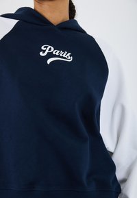 Felpa con cappuccio navy e bianca con una grande grafica "Paris" in bianco corsivo. Presenta una texture morbida e maniche raglan.