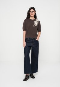 Pull en tricot côtelé marron à manches courtes, orné d'un motif floral blanc sur l'épaule. Associé à un jean denim foncé à jambes larges et des chaussures à talons.