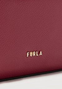 Κοντινό πλάνο υφής από δερμάτινη επιφάνεια μπορντό με μικρά χρυσά γράμματα της επωνυμίας Furla στο κέντρο κοντά στο κάτω άκρο.
