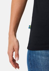 Black t-shirt with short sleeves, featuring a small blue and yellow Swedish flag tag at the hem, paired with blue denim jeans.