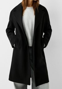 Svart kappa i ullblandning med slagkrage, dubbelknäppt framstycke och sidofickor. Bärs över en grå tröja och jeans.