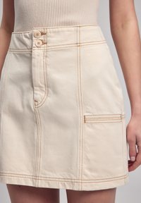 Helle beige Baumwollrock mit glatter Textur, ausgestattet mit zwei Knöpfen an der Taille, kontrastierendem orangefarbenem Nähen und Seitentaschen.
