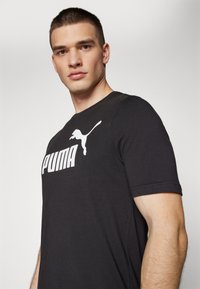 Melna īsām piedurknēm T-kreklā, izgatavots no kokvilnas, priekšpusē atrodas liels balts 'PUMA' logos un pumā siluets. Gluda tekstūra.