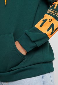Grøn sweatshirt med orange grafik på ærmet, blød tekstur og en forlomme. Ribkanter på ærmer og linning.