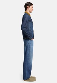 Giubbotto di jeans con colletto senape, corpo blu scuro e tasche laterali. Indossato con jeans a gamba larga blu chiaro e zoccoli beige. Vista di profilo.
