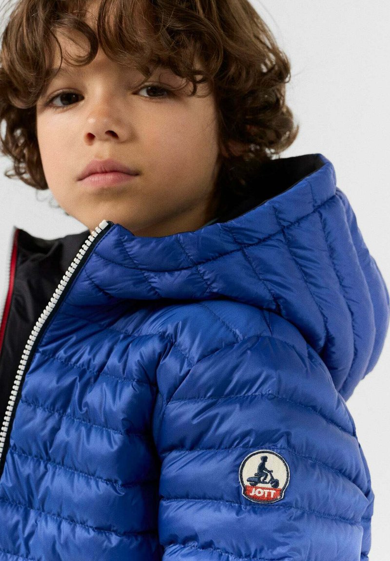 Giacca a vento blu con cappuccio, caratterizzata da una texture trapuntata e un patch rotondo sul manica. Chiusura frontale con zip e rivestimento interno a contrasto.