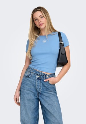 Young woman wearing a light blue short-sleeve top with a shell design, high-waisted blue jeans, and a black shoulder bag.