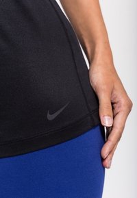 Černý atletický top s mírně zakřiveným lemem a decentním šedým logem Nike. Doplňuje ho modré legíny z hladké, pružné látky.