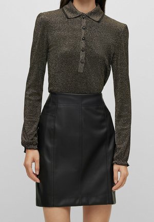 Frau trägt ein metallisch glänzendes Langarmshirt mit Kragen und einen hochgeschnittenen schwarzen Lederrock, steht vor einem schlichten Hintergrund.