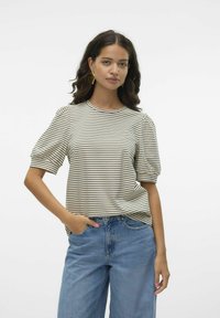 Γυναίκα με σκούρα κυματιστά μαλλιά που φοράει χρυσά σκουλαρίκια σε σχήμα βεντάλιας, ριγέ μπεζ κοντομάνικη μπλούζα και μπλε ψηλόμεσα τζιν, ποζάροντας με το χέρι στην τσέπη.