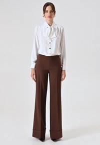 Blouse blanche avec un nœud papillon et fermeture à boutons, associée à un pantalon large taille haute marron avec des poignets structurés.