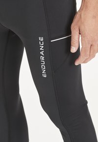 Schwarze Sportleggings mit enger Passform, ausgestattet mit einem weißen Logo "ENDURANCE" und einer kontrastierenden weißen Naht in der Nähe der Tasche.