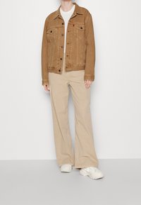 Chaqueta de mezclilla marrón con dos bolsillos en el pecho, botonada al frente y con cuello. Combinada con pantalones anchos de color beige claro y zapatillas blancas.