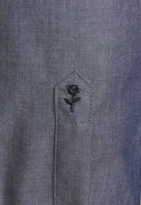 Detail blízkého záběru na tmavě šedou látku se malou černou vyšívanou růží na šitém, šikmém klopně kapsy.