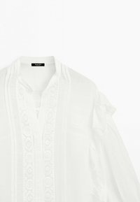 Witte blouse met verticale kanten details en geplooide afwerking langs de voorkant en mouwen, met een kraagloze halslijn.