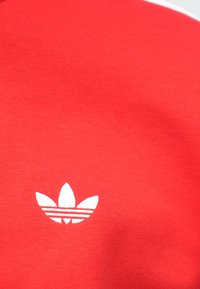 Biele logo Adidas trefoil umiestnené v strede na jasne červenom textilnom pozadí.