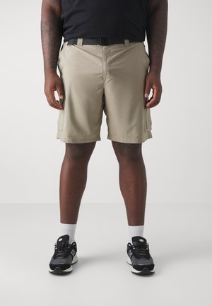 Man wearing beige cargo shorts, black belt, black sneakers with white soles, and white socks standing against a plain light background.