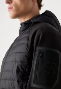 Schwarz gepolsterte Jacke mit glatter Oberfläche, ausgestattet mit einer Kapuze und einer Reißverschlusstasche am linken Ärmel. Logo im Taschenbereich ist deutlich sichtbar.