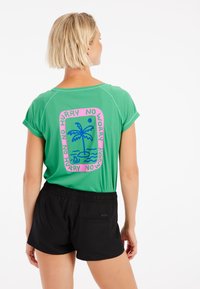 Grön kortärmad skjorta med en färgglad grafik av en palm och text på ryggen, parat med svarta shorts med elastisk midja.