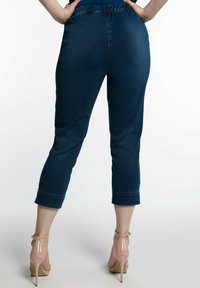 Pantalones recortados de denim azul oscuro con una cintura elástica, que presentan una textura suave y dobladillos en el extremo. Llevados con zapatos de tacón alto en color nude.