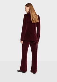 Baršunasti burgundski odijelo, koje se sastoji od krojenog sakoa i hlača širokih nogavica. Tkanina ima meku teksturu i ravnu pozadinu s minimalnim detaljima.