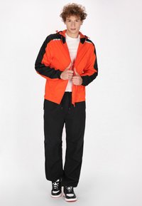 Orange vindjakke med sorte detaljer, udstyret med hætte og front lynlås. Sorte løse bukser og sort-hvide sneakers.