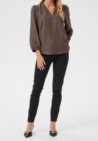 Mujer vestida con blusa marrón de escote en V y mangas abullonadas, pantalones ajustados negros y mules de tacón negro con punta de aguja, de pie frente a un fondo blanco.