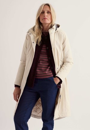 Femme aux longs cheveux blonds portant un manteau crème, un top rayé bordeaux, un cardigan bordeaux et un pantalon bleu, debout avec une main dans la poche.