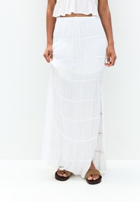 Jupe maxi plissée blanche avec des couches superposées et une taille à cordon. Le tissu est léger et fluide, avec un détail de fente sur le côté.